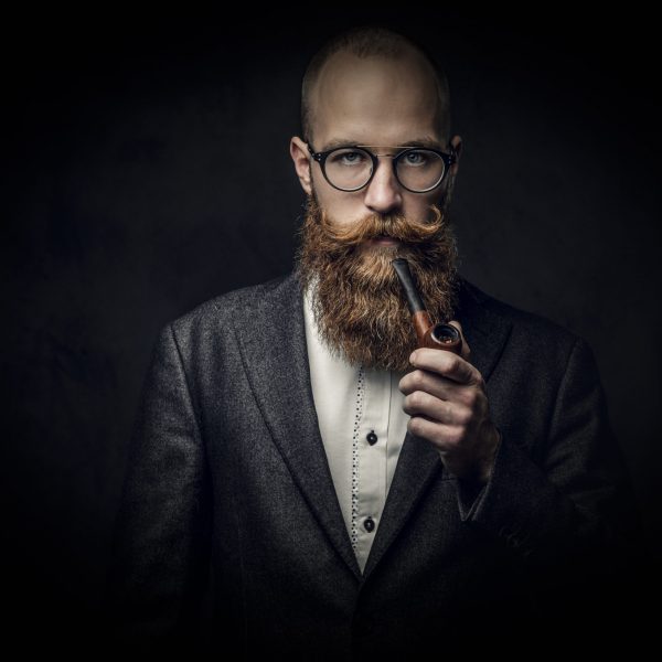 Portrait of bearded redhead English male smoking pipe over grey background.
